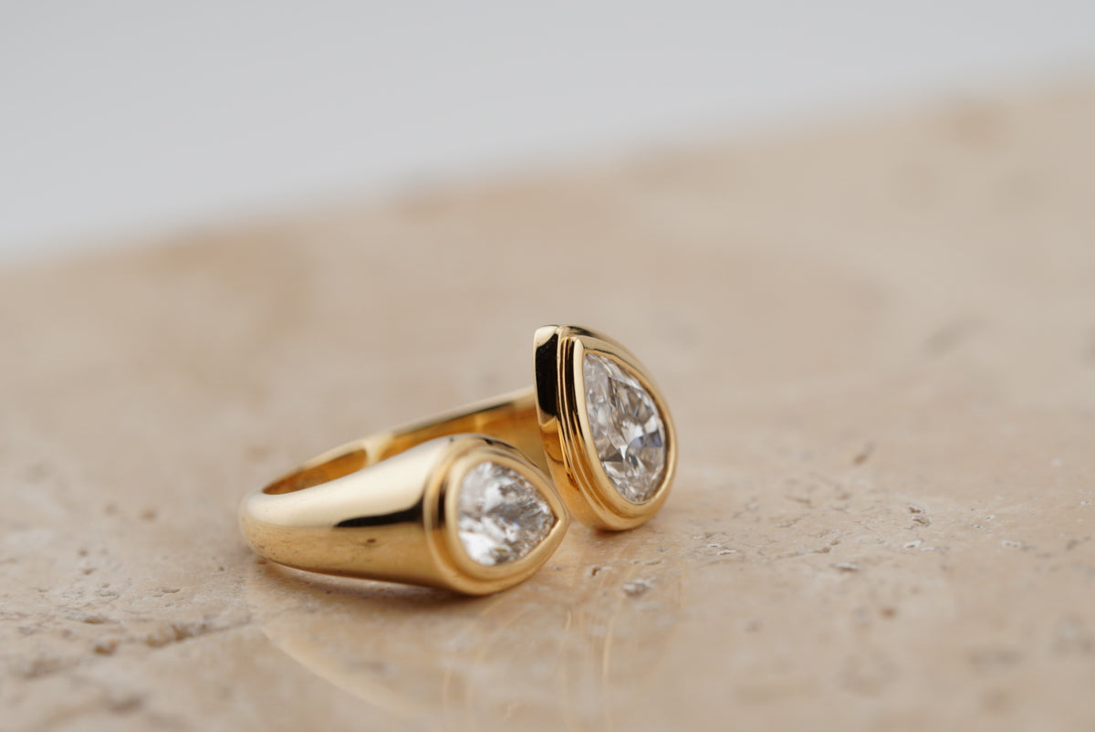 Nora Double Pear Ring in 18K yellow gold, two bezel-set pear-cut diamonds, on travertine